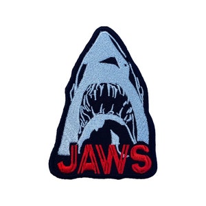 Op de afbeelding: Een geborduurde patch met een open haaienbek in blauw en zwart, met het woord "JAWS" in rode letters.