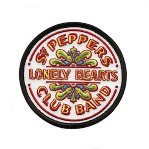 Può includere: Un cerotto rotondo bianco con bordo nero. Il cerotto presenta il testo "Sgt. Pepper's Lonely Hearts Club Band" in lettere rosse e gialle, circondato da un design floreale stilizzato in verde, giallo, blu e rosso.