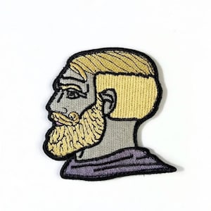 Peut inclure: Écusson brodé représentant le profil d'un homme avec une mâchoire carrée, des cheveux blonds et une barbe fournie. L'écusson présente un visage et un cou gris, un col violet et une bordure noire. Le design est stylisé.