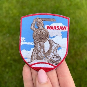 May include: Embroidered patch depicting the Warsaw coat of arms, a mermaid with a sword and shield, set against a blue sky with clouds. The word "WARSAW" is in red. The patch has a red border.