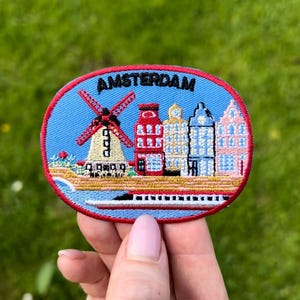 May include: Embroidered patch featuring the word "AMSTERDAM" above a scene with a windmill, canal, and colorful buildings. The patch has a blue background with a red border, showcasing Dutch architecture.