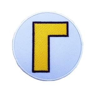 Op de afbeelding: Een witte ronde patch met een gele letter "L" in het midden. De letter is omrand met zwart.