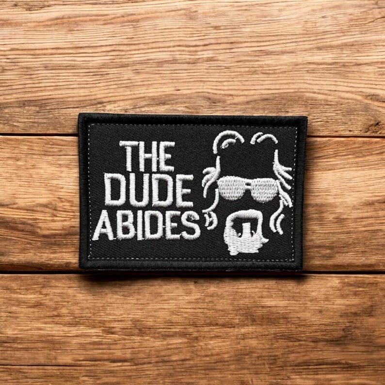 Peut inclure: Patch brod&eacute; noir avec du texte blanc qui dit "The Dude Abides" et une silhouette blanche d'un homme avec des cheveux longs, des lunettes de soleil et une barbe.
