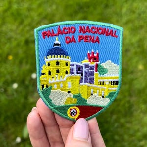 Könnte beinhalten: Gestickter Aufnäher mit dem Palácio Nacional da Pena in Portugal. Der Aufnäher hat eine Schildform mit blauem Himmel und einer farbenfrohen Darstellung des Palastes. Der Text oben lautet "PALÁCIO NACIONAL DA PENA".
