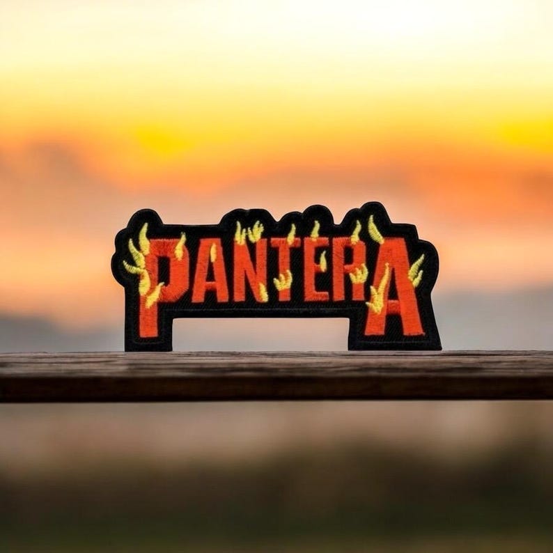 K&ouml;nnte beinhalten: Ein roter und schwarzer gestickter Patch mit dem Wort "PANTERA" in Flammen. Der Patch befindet sich auf einer Holzoberfl&auml;che.