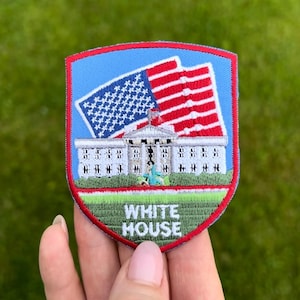 Peut inclure: Écusson brodé représentant la Maison Blanche et le drapeau américain. L'écusson a un fond bleu avec une bordure rouge. Les mots "WHITE HOUSE" sont brodés en blanc.