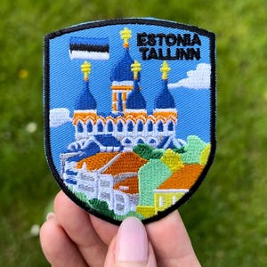 Könnte beinhalten: Gestickter Aufnäher mit einem Schilddesign und dem Text "ESTONIA TALLINN". Der Aufnäher zeigt eine Stadtlandschaft mit blau-bedachten Gebäuden, orangefarbenen Dächern und der estnischen Flagge. Der Aufnäher wird vor einem grünen Hintergrund gehalten.