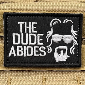 Op de afbeelding: Een zwarte patch met witte geborduurde tekst die "The Dude Abides" luidt. De patch heeft een witte geborduurde silhouet van een man met lang haar, een zonnebril en een baard.