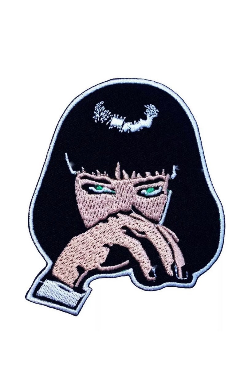 May include: Embroidered iron-on patch featuring a black and white illustration of a woman with green eyes, covering her mouth with her hand.