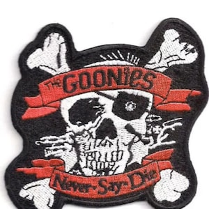 May include: Black and white embroidered patch featuring a skull and crossbones. The skull has a red bandana with the text "The Goonies" and a red ribbon with the text "Never Say Die".