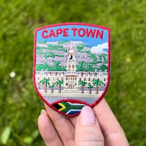 May include: Embroidered patch featuring a detailed depiction of Cape Town, South Africa, with the text "CAPE TOWN" at the top. The design includes a building, mountains, palm trees, and the South African flag at the bottom. The patch has a red border.