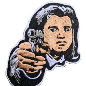 Puede incluir: Parche bordado con una ilustración en blanco y negro de una persona sosteniendo un arma de fuego, con la cara marrón y el pelo negro.