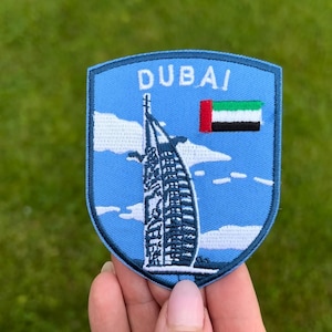 Op de afbeelding: Een blauwe geborduurde patch met het woord "DUBAI" bovenaan. De patch toont het Burj Al Arab hotel, een wit gebouw met een zeilachtig ontwerp, tegen een blauwe lucht. De vlag van de VAE staat rechts.