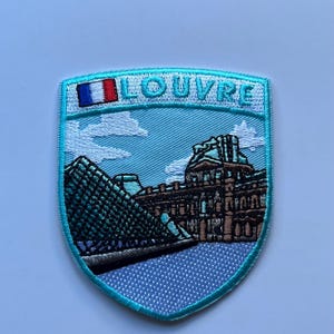 Può includere: Toppa ricamata raffigurante il Museo del Louvre a Parigi. La toppa ha la forma di uno scudo con un bordo azzurro. Il disegno comprende la piramide e l'edificio del museo, la parola "LOUVRE" e una piccola bandiera francese.
