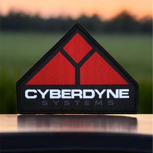 May include: A black and red embroidered patch with the text "CYBERDYNE SYSTEMS" in white. The patch features a stylized triangle with three smaller triangles inside.