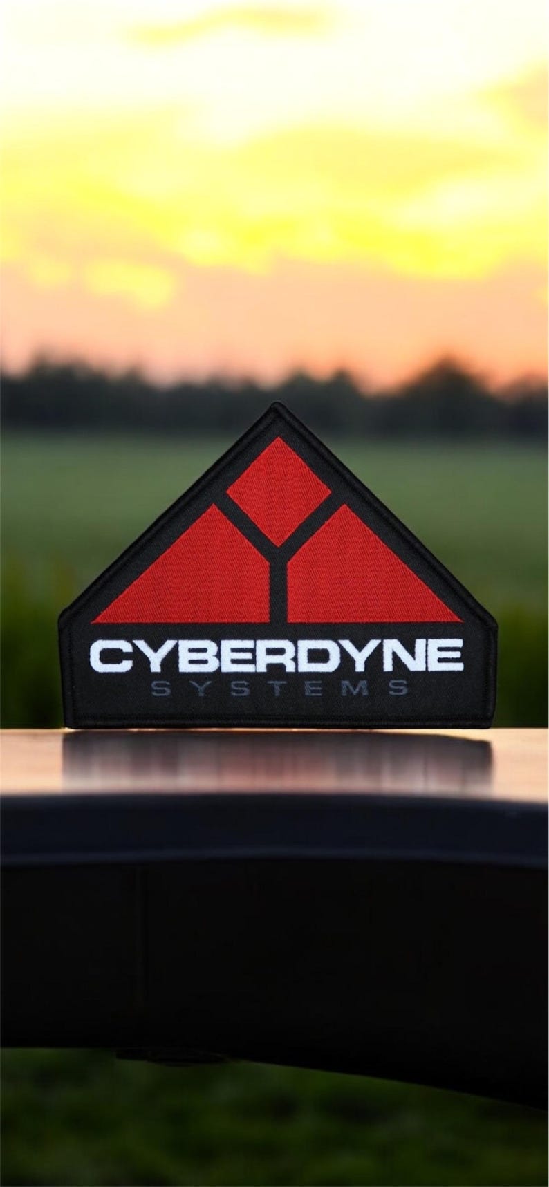 May include: A black and red embroidered patch with the text "CYBERDYNE SYSTEMS" in white. The patch features a stylized triangle with three smaller triangles inside.