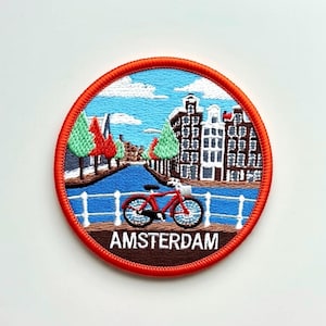 Può includere: Toppa rotonda ricamata raffigurante una scena del canale di Amsterdam. Una bicicletta rossa è su un ponte, con edifici, alberi e un cielo blu nuvoloso. La parola "AMSTERDAM" è ricamata in bianco su sfondo marrone, incorniciata da un bordo rosso.