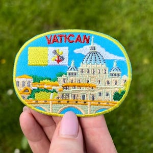 Può includere: Toppa ricamata raffigurante la Città del Vaticano. La toppa ha un bordo ovale blu con la scritta "VATICAN" in alto. Il disegno comprende la bandiera vaticana, la Basilica di San Pietro e un ponte, il tutto in colori vivaci.