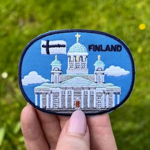 Pode incluir: Um patch oval com uma borda azul marinho e um fundo azul, representando um edifício com uma cruz, a bandeira finlandesa e a palavra "FINLAND". O edifício é branco com um telhado verde e tem nuvens brancas em ambos os lados. O patch é segurado por uma pessoa.