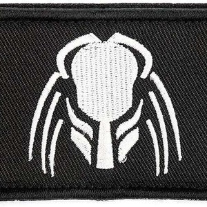 May include: Black rectangular patch featuring a white embroidered alien face design. The patch has a black border and is likely an emblem or insignia.