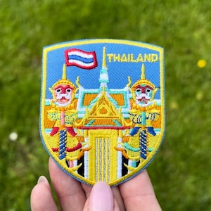 Puede incluir: Parche bordado con un diseño de escudo que representa un templo tailandés y dos figuras de guardianes. El parche tiene un fondo azul con la palabra "THAILAND" y una bandera tailandesa. El parche es amarillo, rojo y azul.