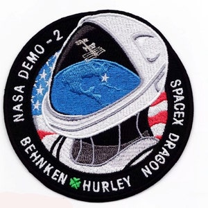 Op de afbeelding: Een ronde, zwart-witte geborduurde patch met de tekst "NASA DEMO-2 BEHNKEN HURLEY SPACEX DRAGON" rond de rand. Het midden van de patch toont een witte astronautenhelm met een blauw en wit beeld van de aarde erin. De astronautenhelm is gedeeltelijk bedekt met een rood-wit-blauwe Amerikaanse vlag.