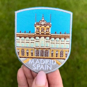 Op de afbeelding: Geborduurd schildvormig embleem met een gedetailleerde illustratie van een gebouw tegen een blauwe lucht. De patch heeft de tekst "MADRID SPAIN" aan de onderkant. Het gebouw is weergegeven in bruin-, beige- en wittinten, wat wijst op een paleis.