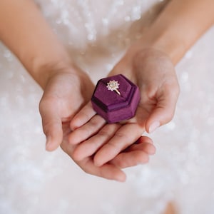 Könnte beinhalten: Eine violette Samt-Ringbox mit einem goldenen Ring im Inneren. Der Ring hat einen großen, klaren Stein in der Mitte, umgeben von kleineren Steinen.