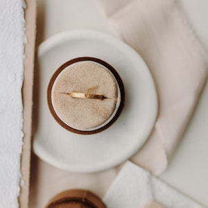 Op de afbeelding: Een gouden ring met een klein hartvormig ontwerp ligt op een beige fluwelen ringdoos. De ringdoos staat op een wit bord. Het bord ligt op een beige oppervlak.