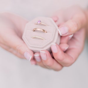 Könnte beinhalten: Eine kleine, hellrosa Ringbox mit drei goldenen Ringen. Der oberste Ring hat einen kleinen rosafarbenen Edelstein. Die Box wird in den Händen einer Person gehalten.