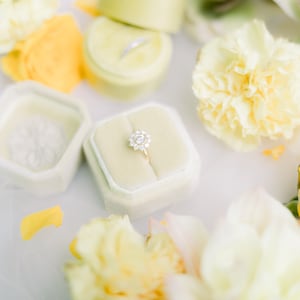 Può includere: Una scatola per anelli bianca con un interno in velluto e un anello di diamanti all'interno. La scatola è circondata da fiori gialli e bianchi.