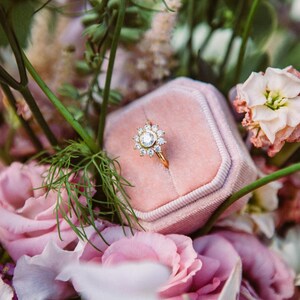 May include: A pink velvet ring box with a gold ring with a large, clear center stone surrounded by smaller stones. The box is surrounded by pink and white flowers.