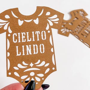 May include: Two brown paper cutouts in the shape of a baby onesie. The text "CIELITO LINDO" is cut out in white. The onesies have decorative cut-out floral designs around the edges. One is held by a hand.