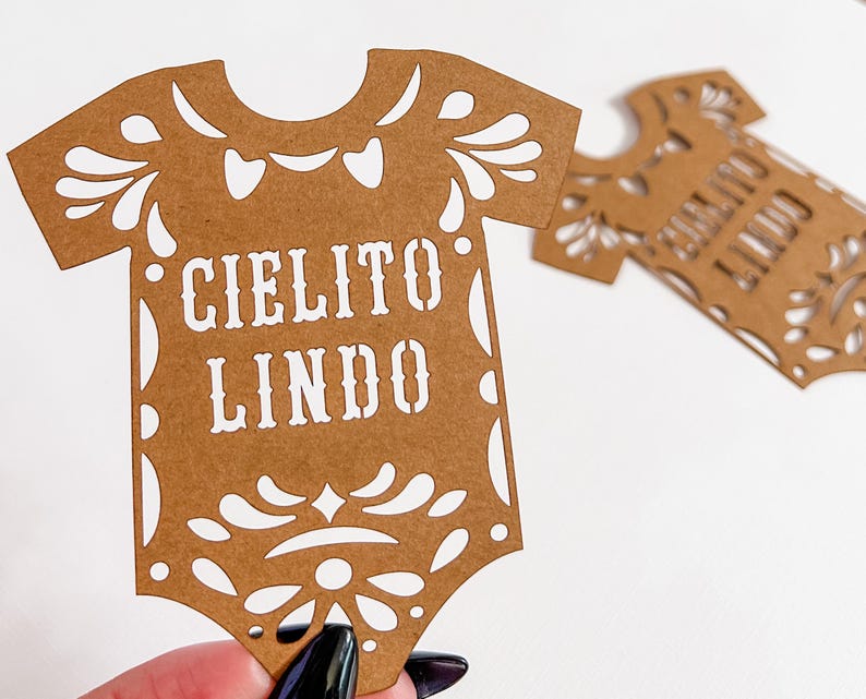 May include: Two brown paper cutouts in the shape of a baby onesie. The text "CIELITO LINDO" is cut out in white. The onesies have decorative cut-out floral designs around the edges. One is held by a hand.