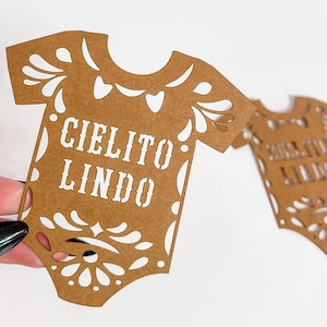 May include: Brown paper cutout in the shape of a baby onesie with the words "CIELITO LINDO" in white. The onesie has decorative cutouts around the edges. Another similar cutout is visible in the background.