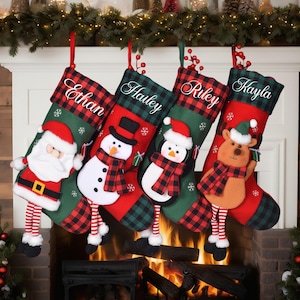 May include: Four Christmas stockings hang above a fireplace. Each stocking is red and green plaid with a name embroidered on the cuff. Santa, snowman, and reindeer figures are attached to the stockings.