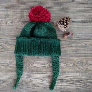 Puede incluir: Un gorro de punto verde con un pompón rojo en la parte superior y orejeras largas. El gorro está sobre una superficie de madera.