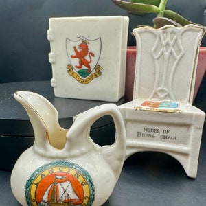 May include: A collection of three small, white ceramic souvenirs. Includes a small pitcher with a sailboat emblem, a book-shaped item with a lion crest, and a chair-shaped item with the text "MODEL OF BURNS CHAIR".