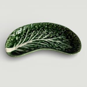 May include: A ceramic leaf-shaped dish, dark green with a white vein pattern, resembling a cabbage leaf. The curved dish is likely for serving food or as a decorative item. The dish's measurements are in centimetres.