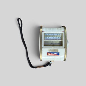May include: A vintage Sixtomat light meter featuring a white rectangular body with gold-toned metal side panels. The device includes a display with numerical scales, a needle indicator, and a black fabric wrist strap attached to the bottom.