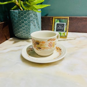 Peut inclure: Une tasse à thé et une soucoupe blanches avec un motif floral composé de fleurs brunes, orange et roses et d'épis de blé. La soucoupe a un bord festonné.