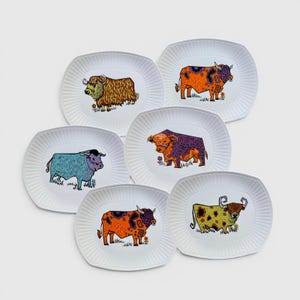 May include: A set of six white oval plates with ribbed edges, each featuring a unique illustration of a bull. The bulls are depicted in various colours including orange, purple, blue, and yellow, with intricate patterns and small floral details.