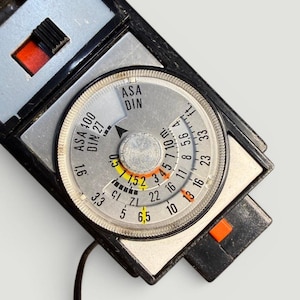 Könnte beinhalten: Nahaufnahme eines Vintage-Belichtungsmessers mit einem kreisförmigen Zifferblatt. Das Zifferblatt zeigt Zahlen, Skalen und den Text "ASA DIN". Der Belichtungsmesser hat ein schwarz-silbernes Gehäuse mit orangefarbenen Akzenten und einem schwarzen Kabel.