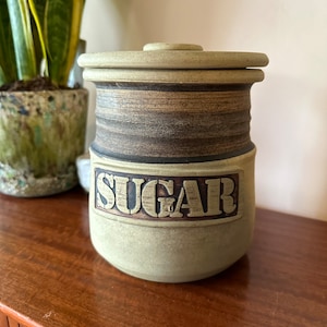 Könnte beinhalten: Eine grüne Keramik-Zuckerdose mit Deckel. Die Dose hat braune Streifen und das Wort "SUGAR" in braunen Buchstaben.