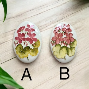 May include: Two white oval ceramic tiles with a raised floral design. The design features pink flowers with green leaves. The tiles are labelled A and B.