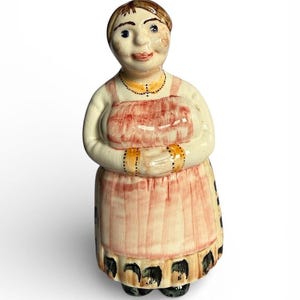May include: A ceramic figurine of a woman wearing a white blouse, pink apron, and black shoes. The apron has a pink and white striped pattern. The figurine has brown hair and is wearing a gold necklace and bracelets.