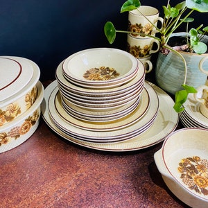 Puede incluir: Un juego de platos de cerámica vintage con estampado floral, que incluye una fuente cubierta, cuencos, platos y tazas de té. Los platos son en tonos marrones, crema y blanco con un diseño floral.