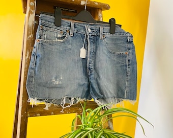 May include: A pair of light blue denim shorts with a frayed hem. The shorts are hanging on a wooden ladder against a yellow wall.