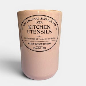 May include: A light pink ceramic kitchen utensil jar with a tapered cylindrical shape. The front features black printed text reading THE ORIGINAL SUFFOLK JAR, KITCHEN UTENSILS, Ideal for Use at Home or on Safari, HENRY WATSON POTTERY ENGLAND Founded 1800.