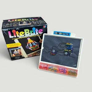 以下が含まれることがあります： 黒い箱と白いフレームのLite-Briteおもちゃセット。箱にはLite-Briteのロゴとカラフルなデザインの画像が描かれています。フレームは、画像を生成するために挿入されたカラフルなペグを備えた暗い画面です。コンパートメントには、さまざまな色のペグが収納されています。